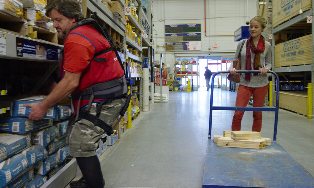 Lowe's Home Improvement associate on sales floor lifting heavy object for female customer while wearing soft robotic exoskeleton for assistance.